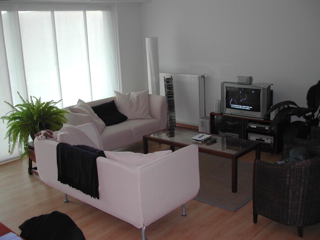 A living room with two white sofas, a glass coffee table, and a TV playing a subtitled program.