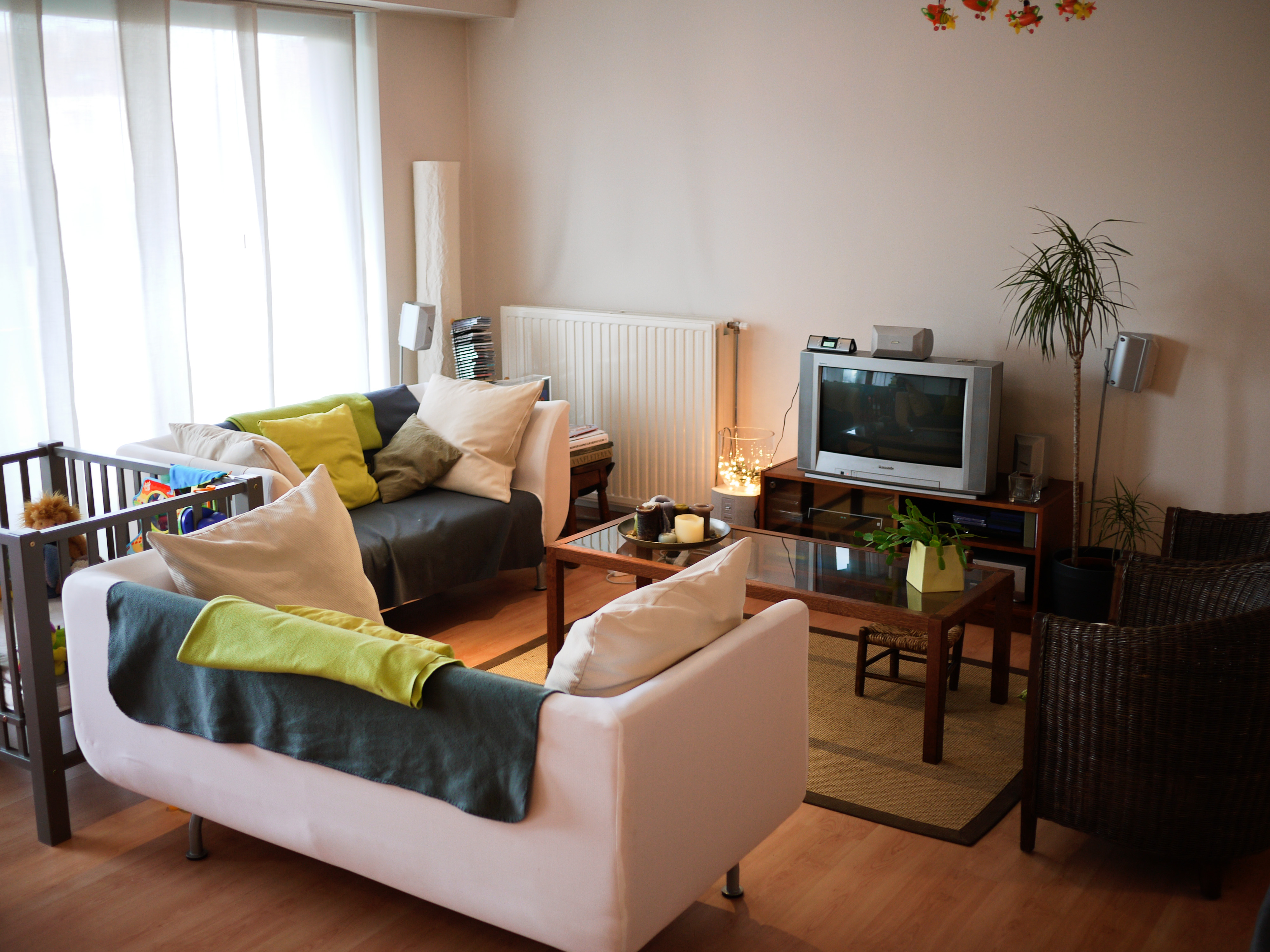 A cozy living room with two sofas, a coffee table, a TV, and a playpen near the window.