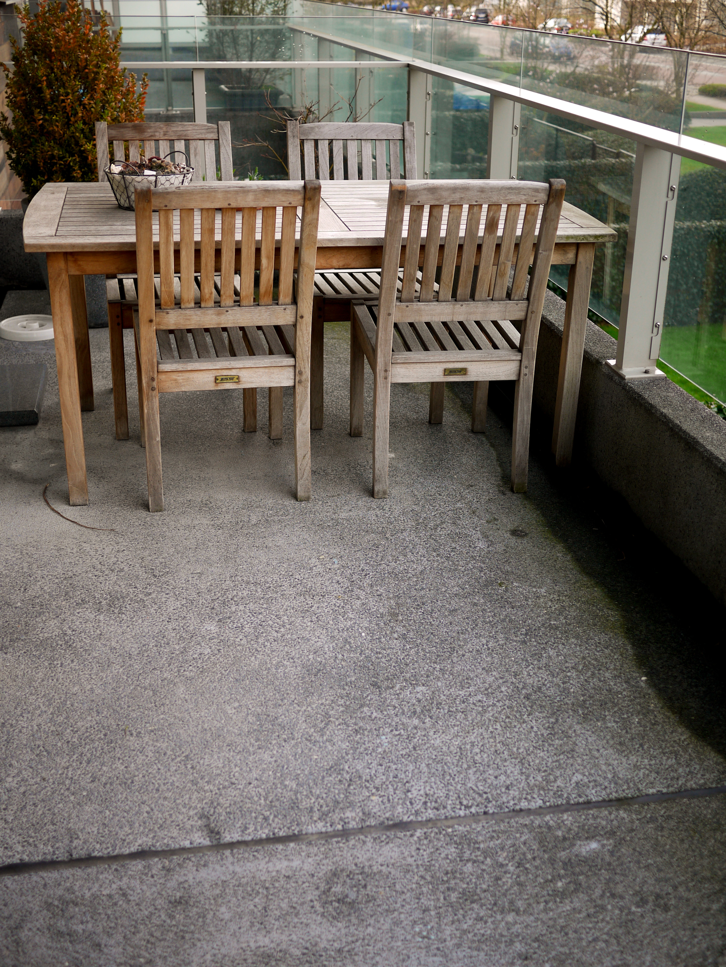 Wooden table with four matching chairs on a concrete balcony with glass railings, overlooking a street and greenery.