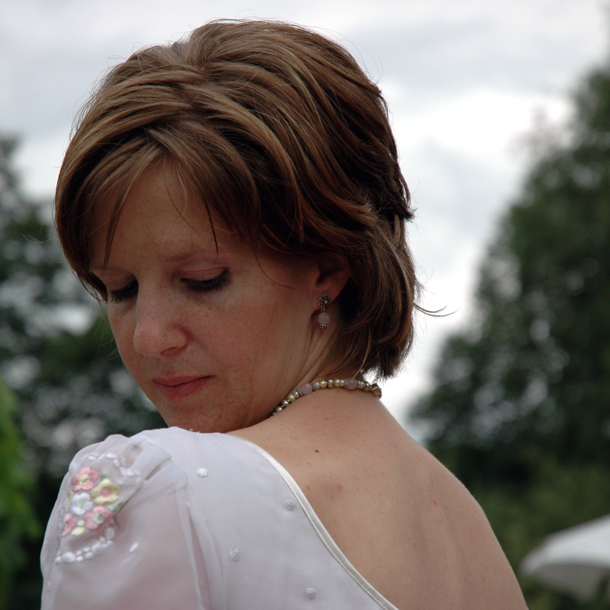 A woman in a white dress with floral details looks down, wearing a pearl necklace and earrings.