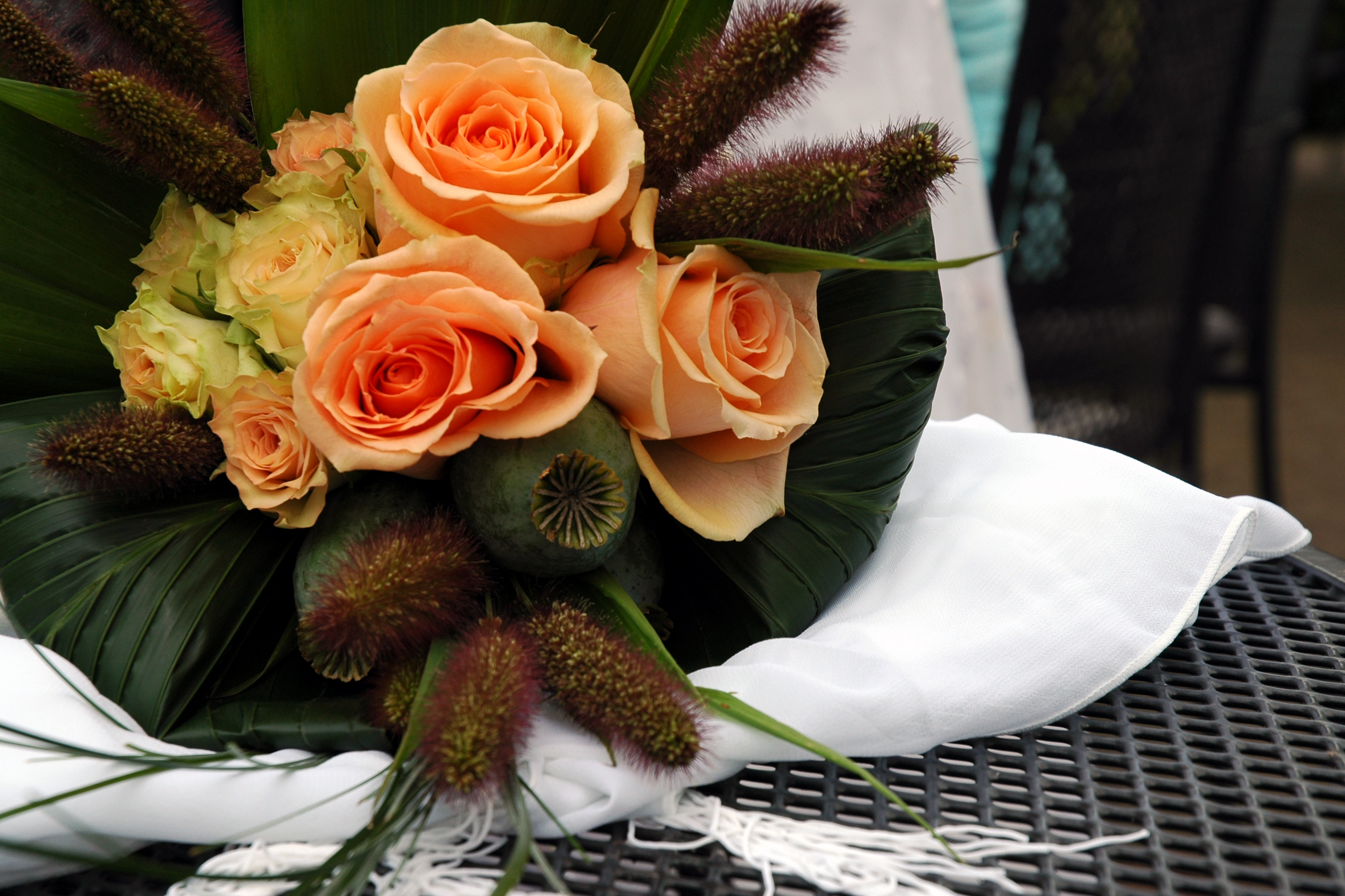 A bouquet of orange and yellow roses with green leaves and decorative plants rests on a white cloth.