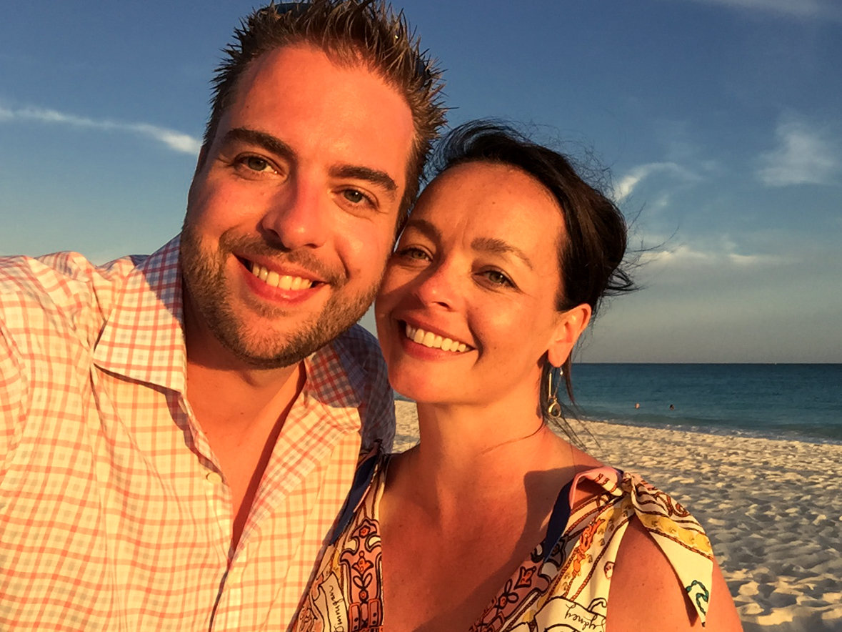A smiling couple takes a close-up selfie on a sandy beach with the ocean in the background.