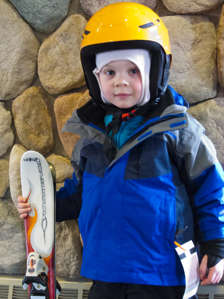 A young child wearing a yellow helmet and blue ski jacket holds skis, ready to go skiing.