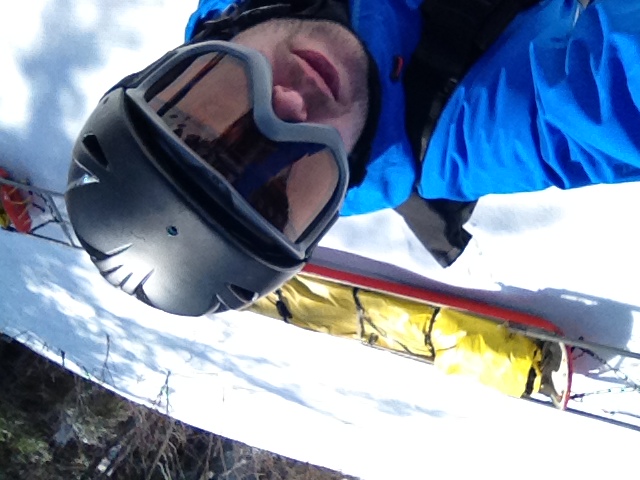 Person in ski gear taking a selfie on snowy ground, with a rescue sled carrying someone in the background.