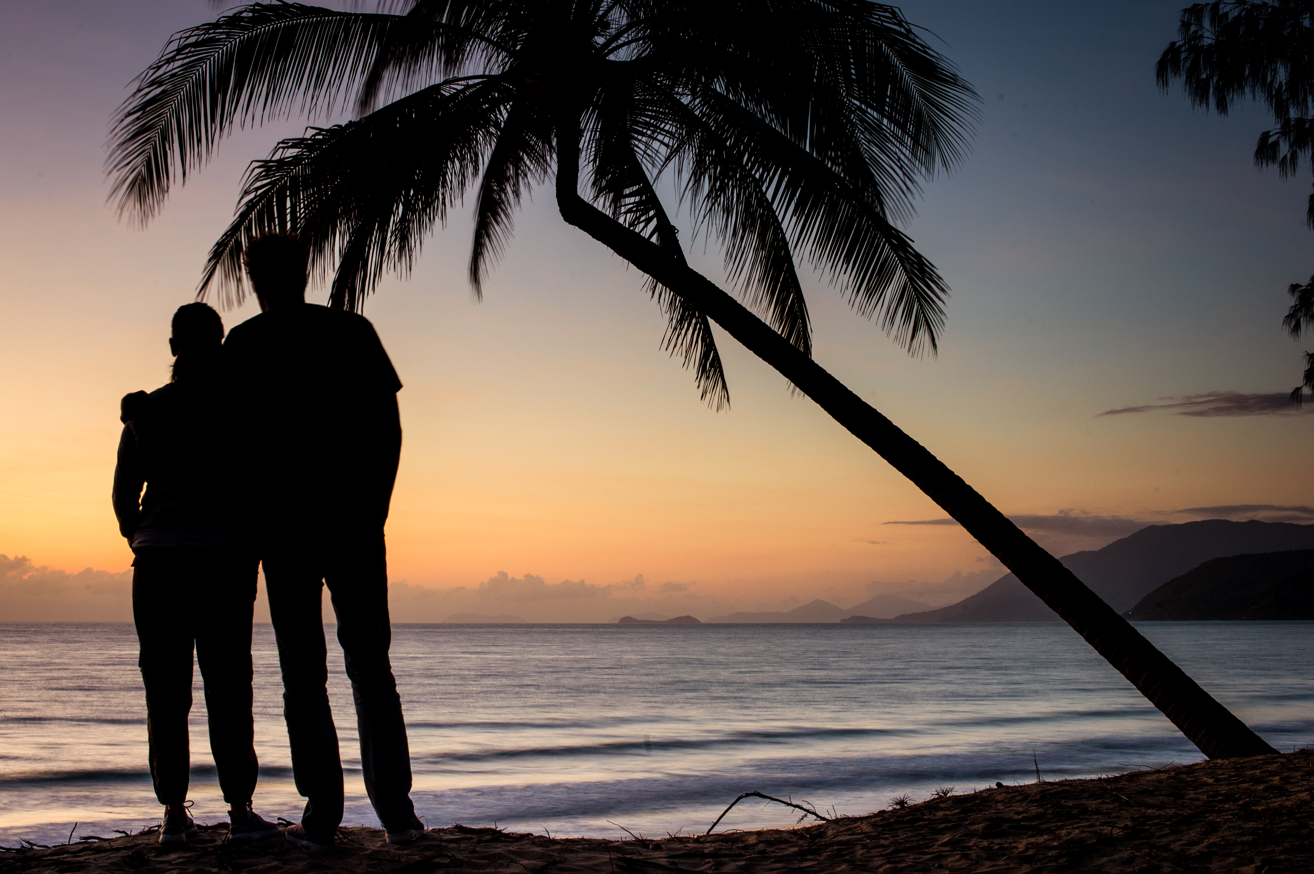 Two people stand close together on a beach, looking at the ocean during sunrise.