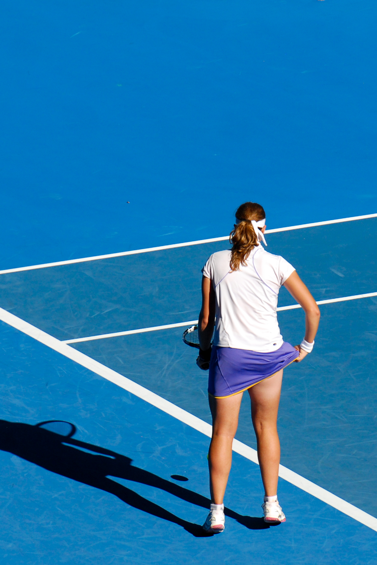 A tennis player stands on the blue court, holding a racket and adjusting her skirt during a match.