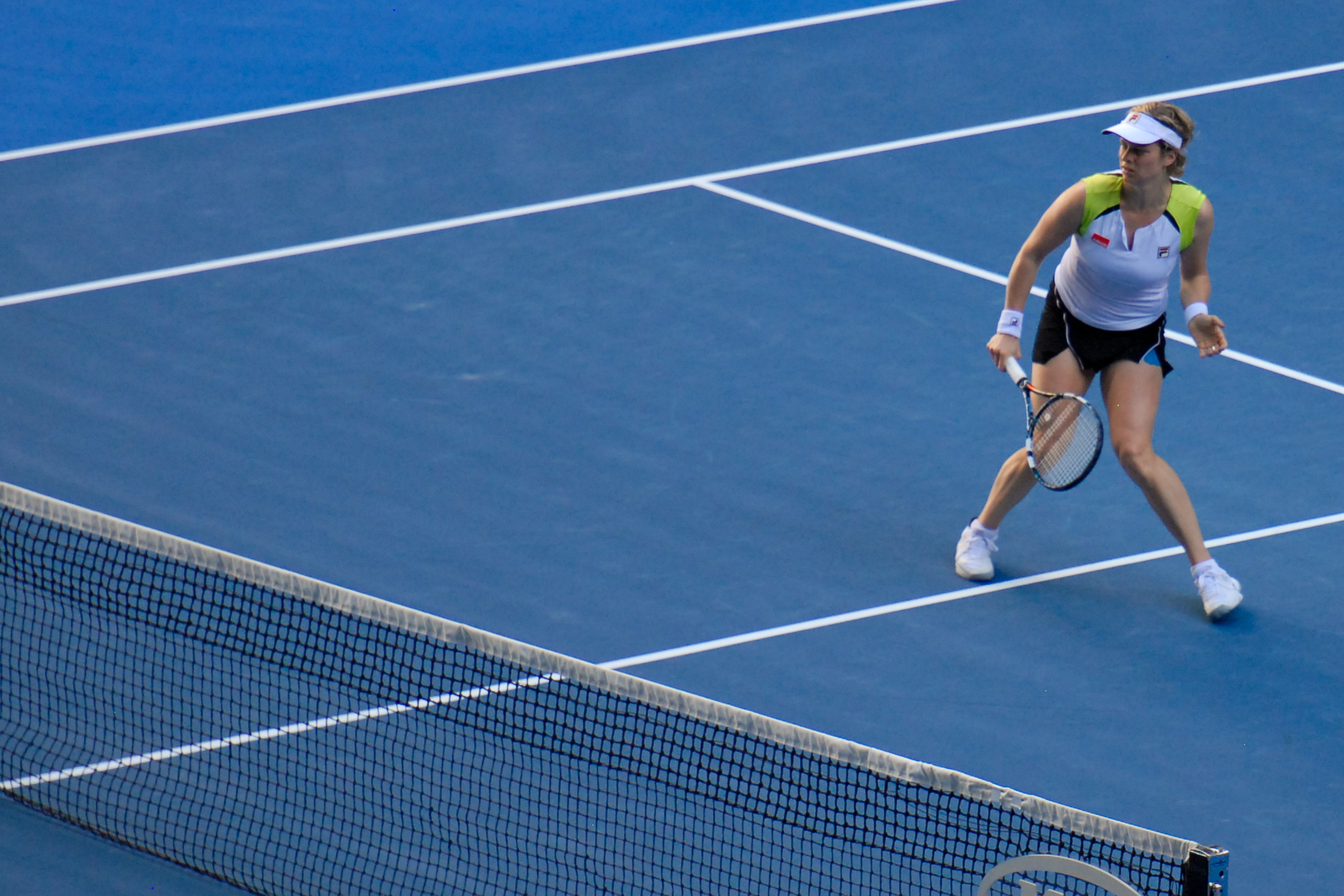 A tennis player prepares to return a shot during a match on a blue hard court.