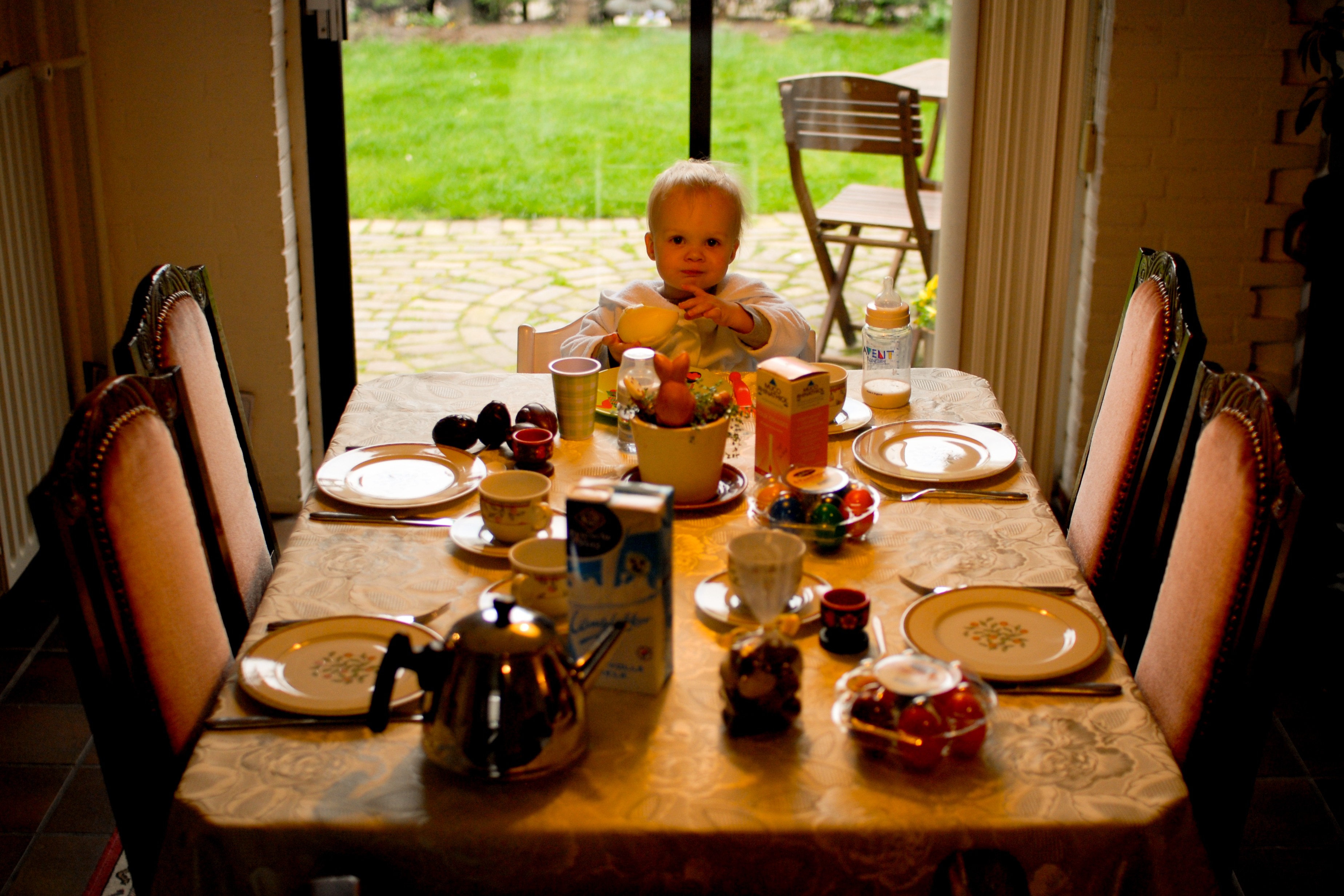 A young child sits at the head of a dining table, holding a bowl, surrounded by breakfast items.