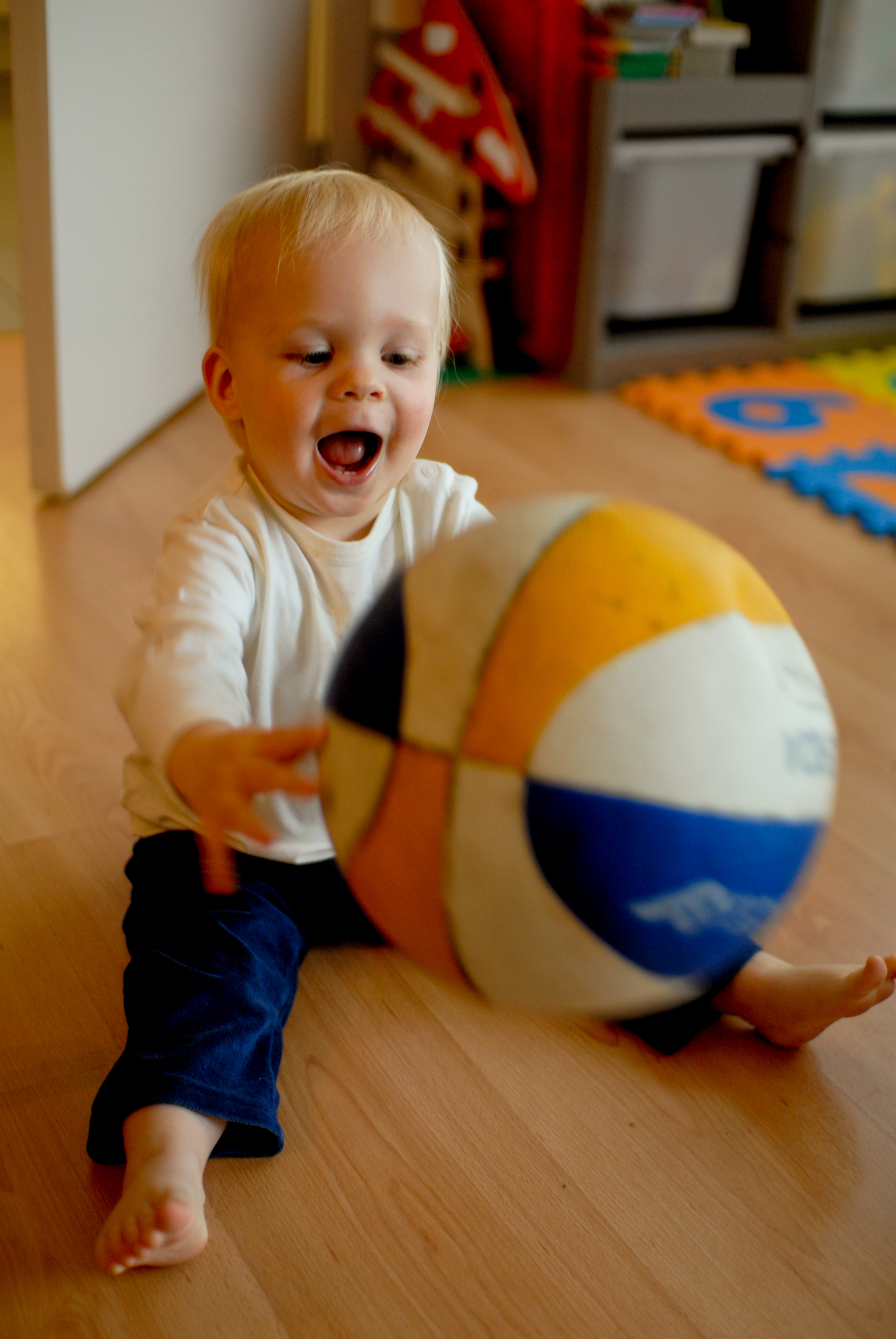 A young child sits on the floor, reaching out with excitement to catch a colorful ball.