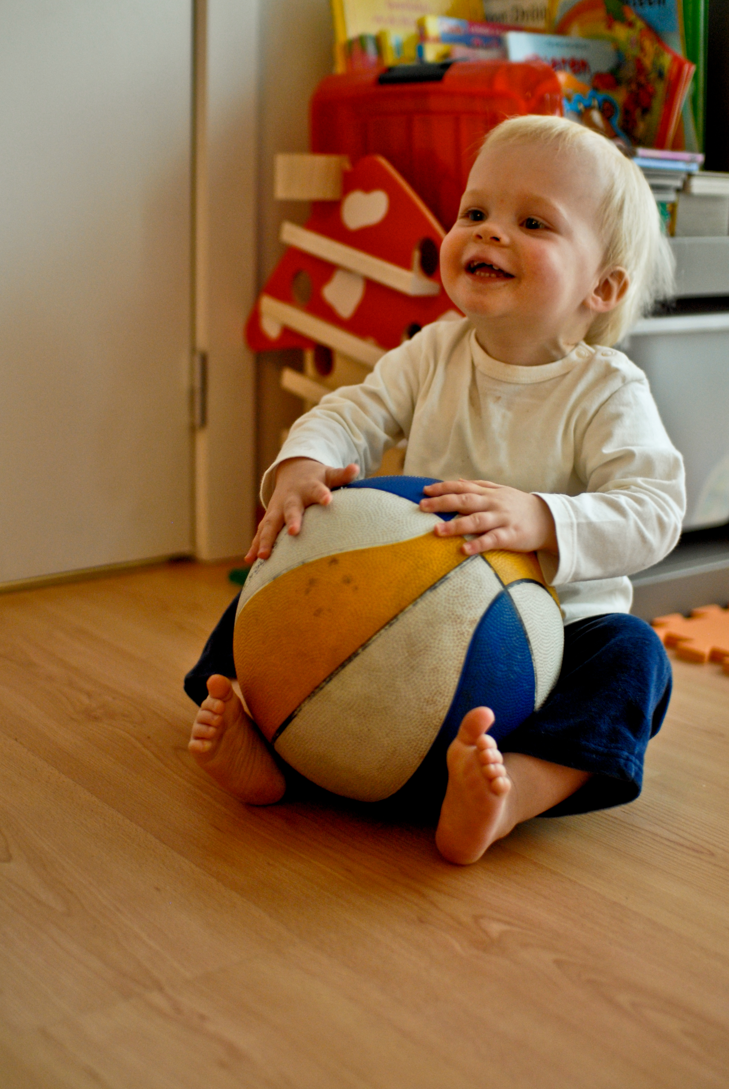 A smiling toddler sits on the floor, holding a large, colorful ball with both hands.