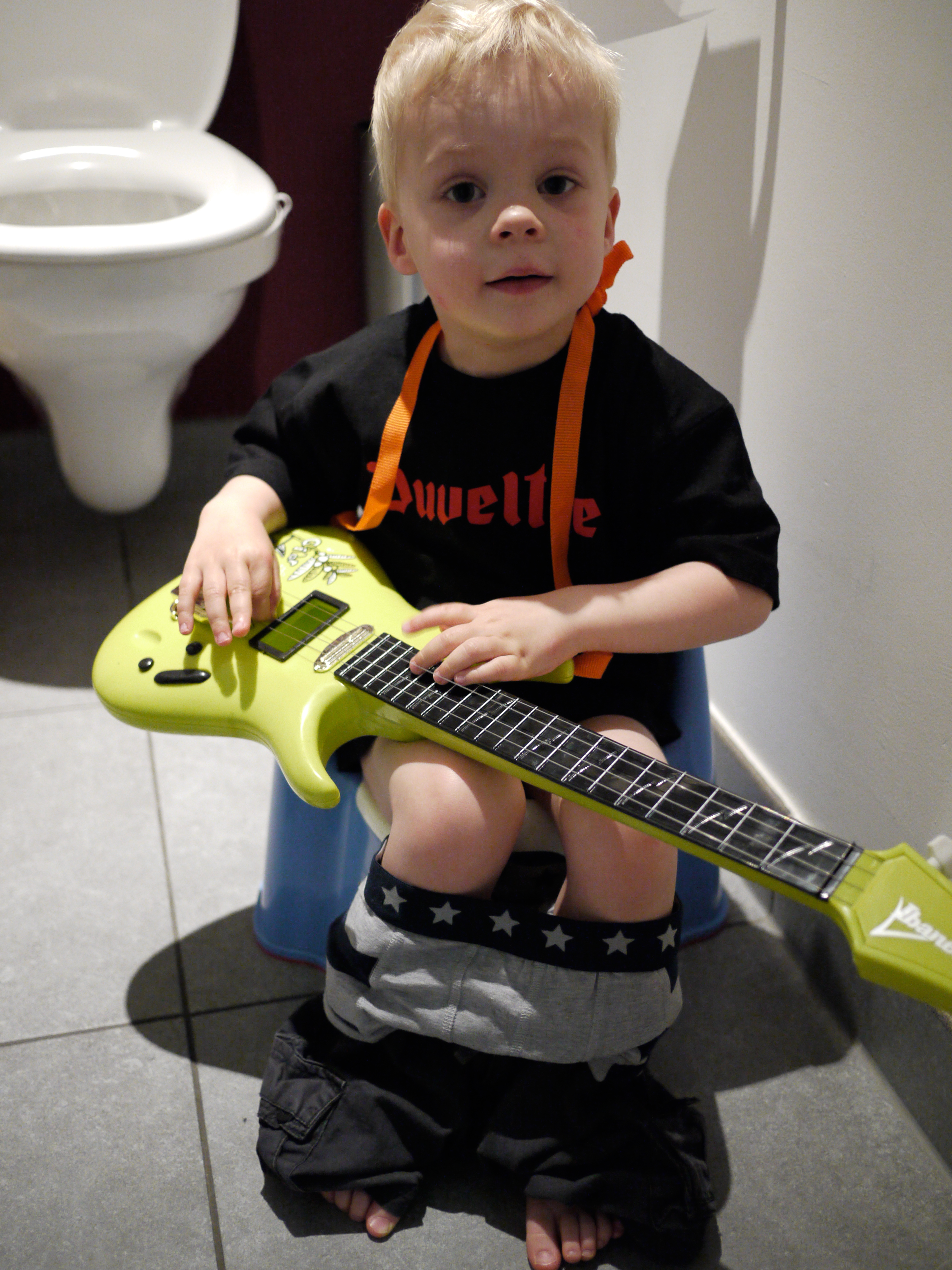 A young child sits on a potty with pants down, holding a green toy guitar and looking at the camera.