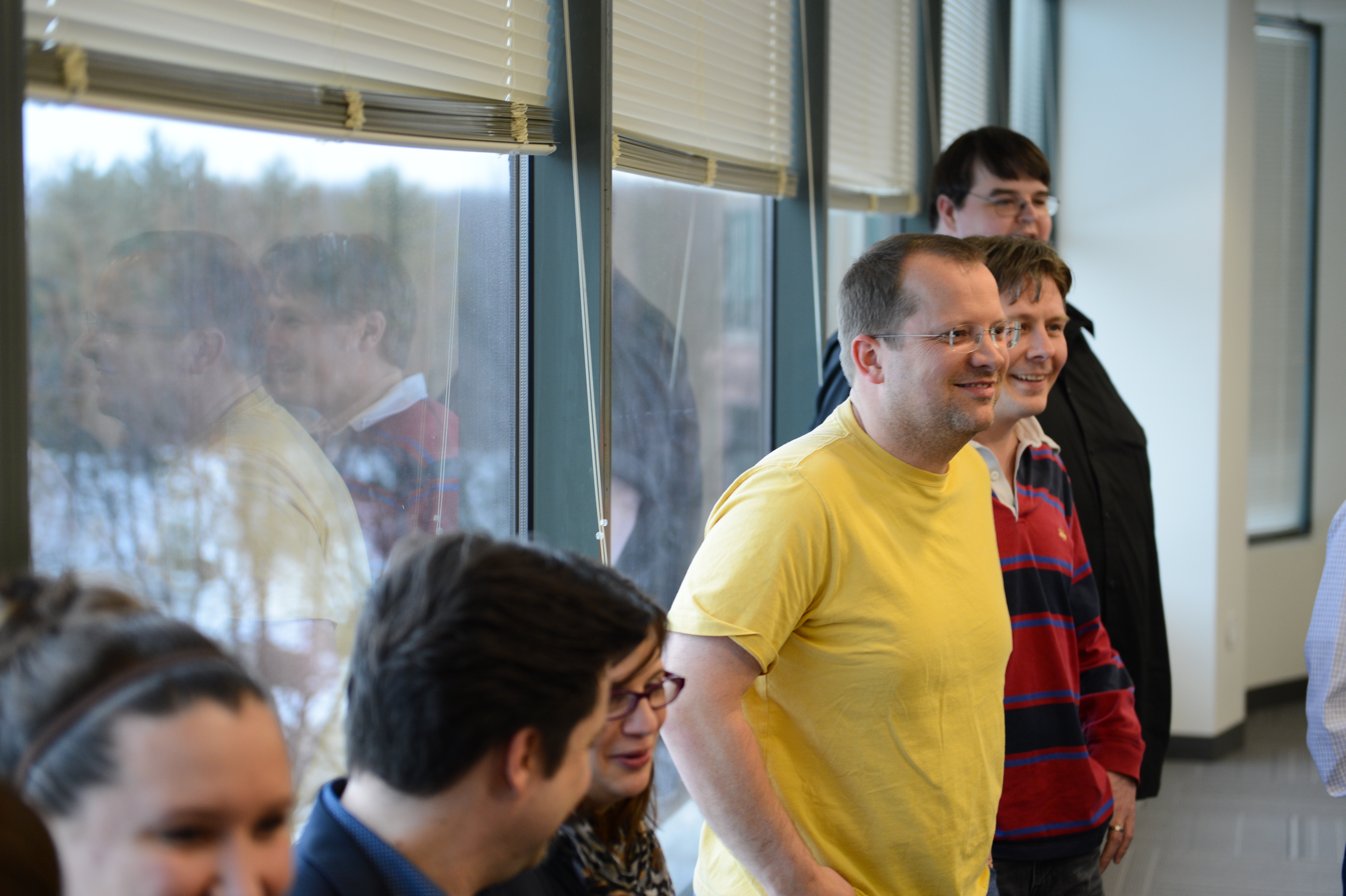 A group of people standing near windows, smiling and engaged in conversation at a baby shower event.