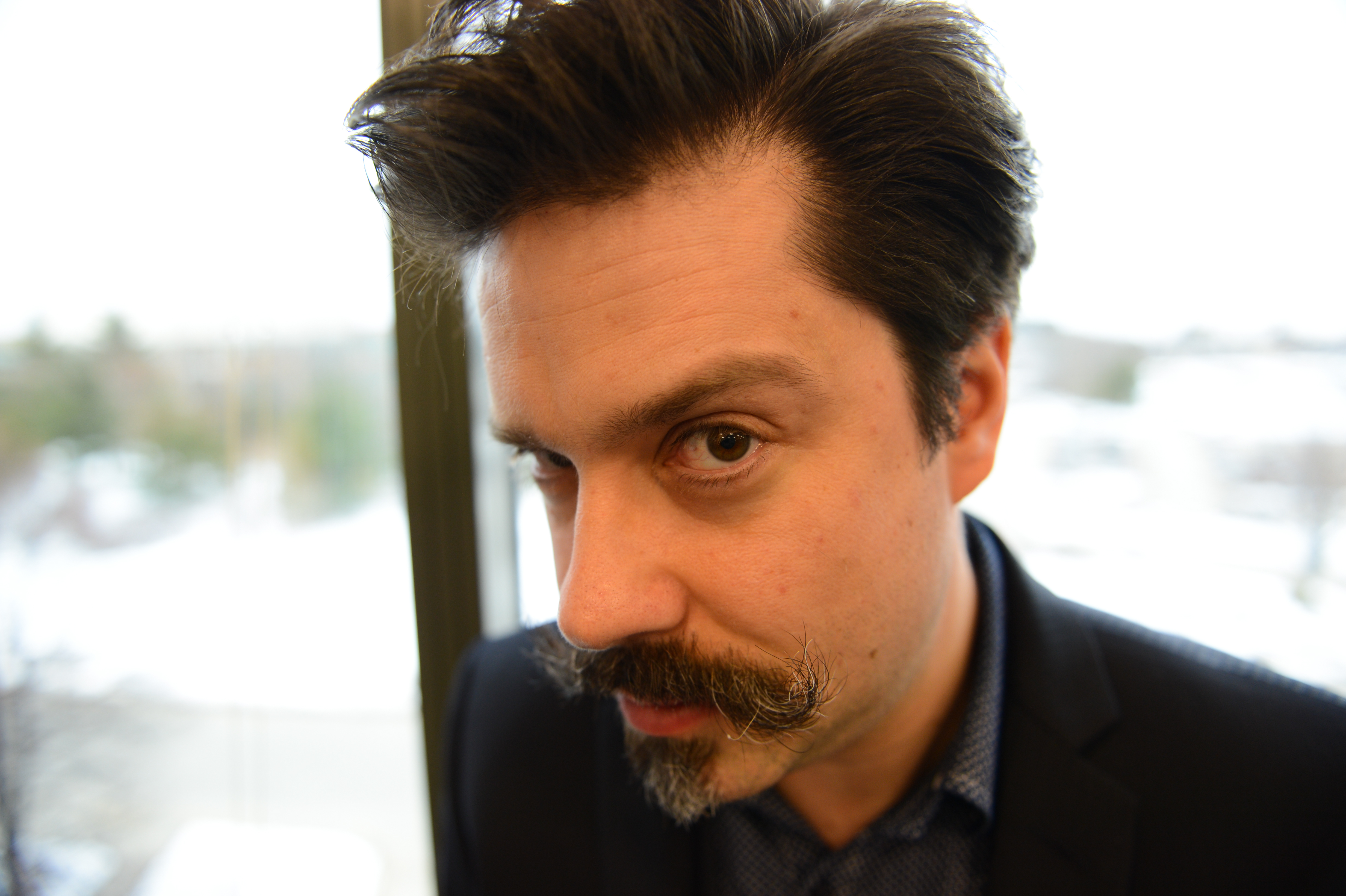 A man with a mustache looks directly at the camera with a curious expression indoors near a window.