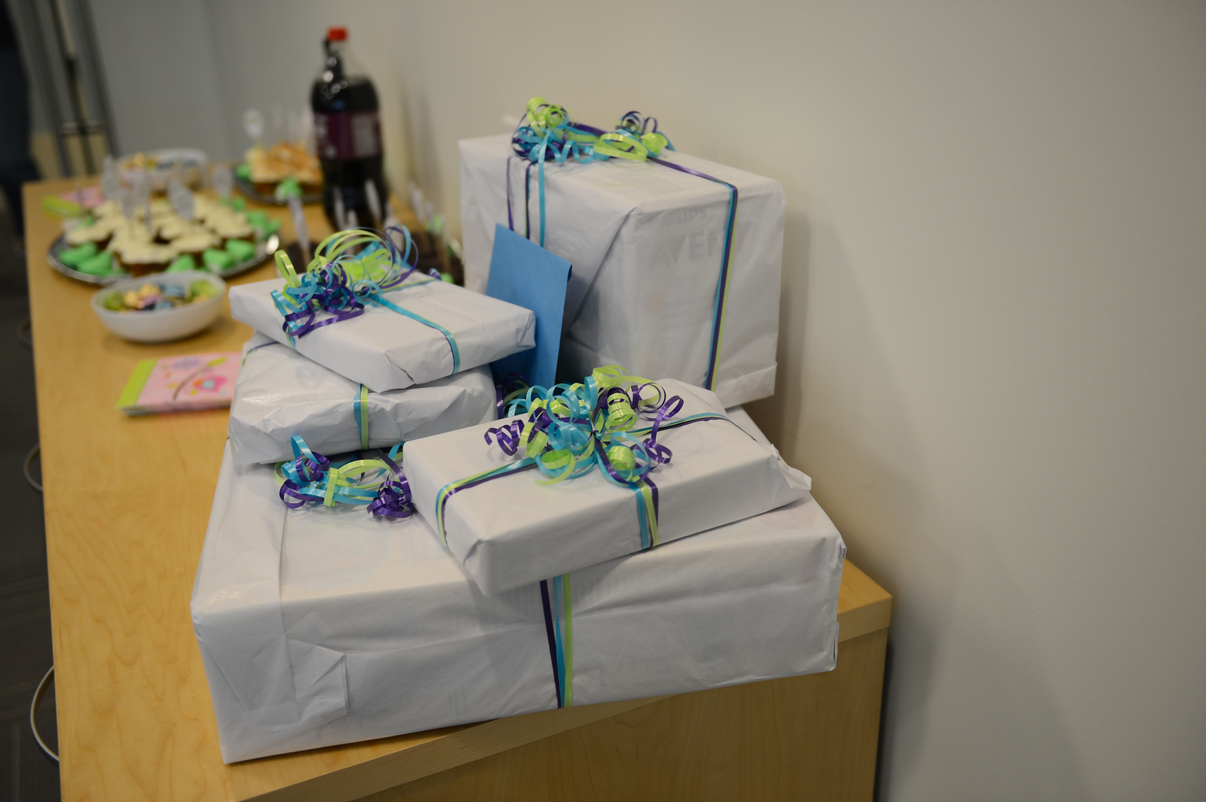 A stack of wrapped gifts with ribbons on a table, with snacks and drinks in the background.