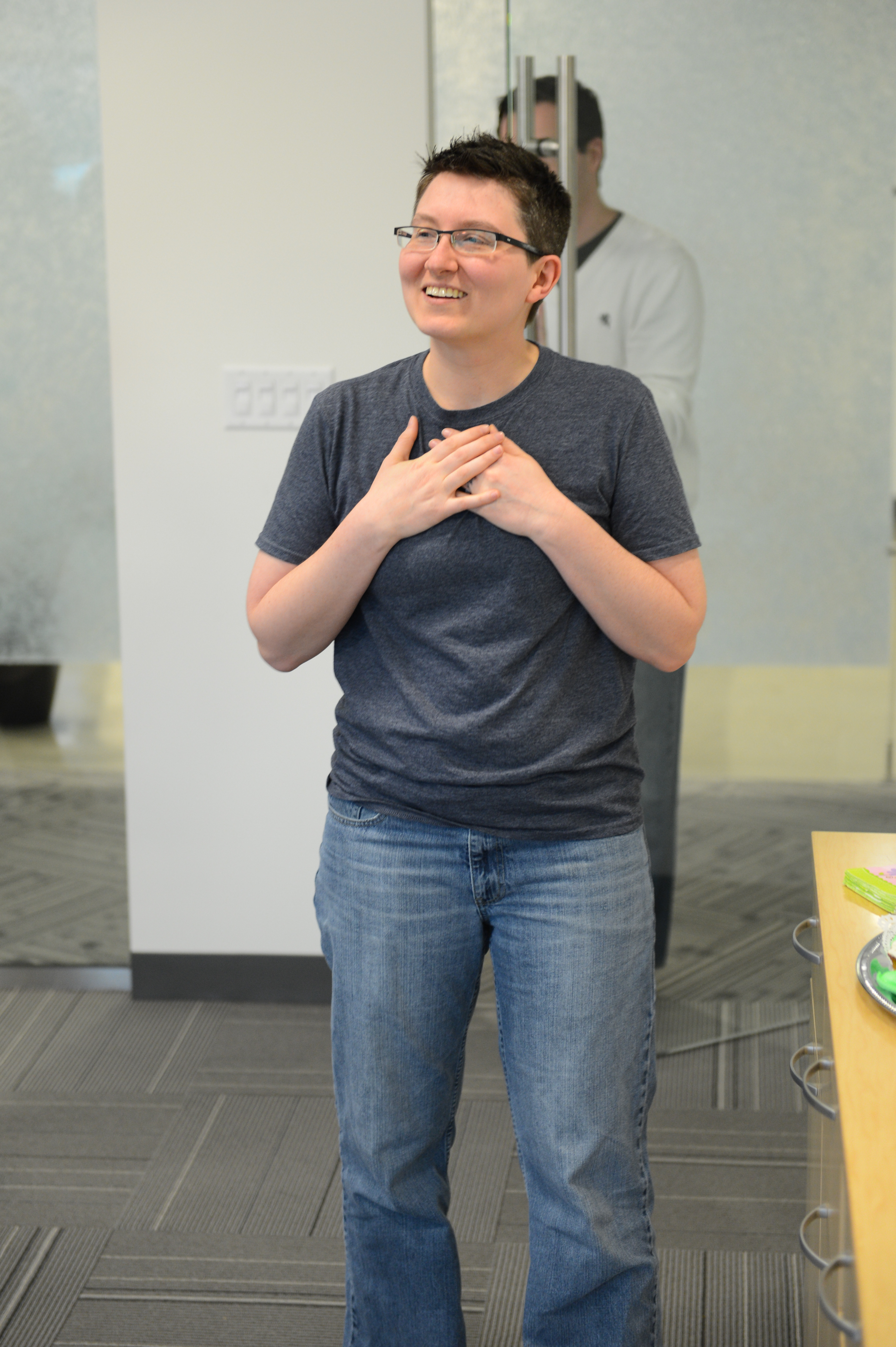 A person in a dark shirt and jeans smiles with hands on their chest, appearing touched or grateful.
