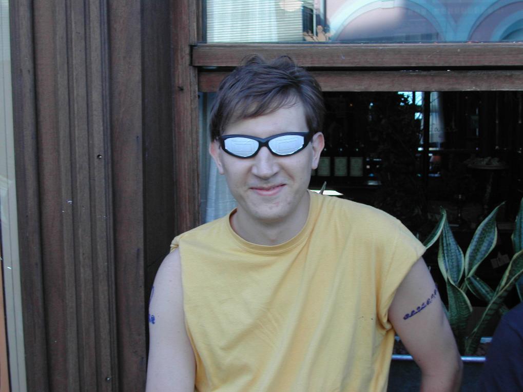 A man in a yellow sleeveless shirt and sunglasses smiles slightly while sitting outside near a wooden structure.