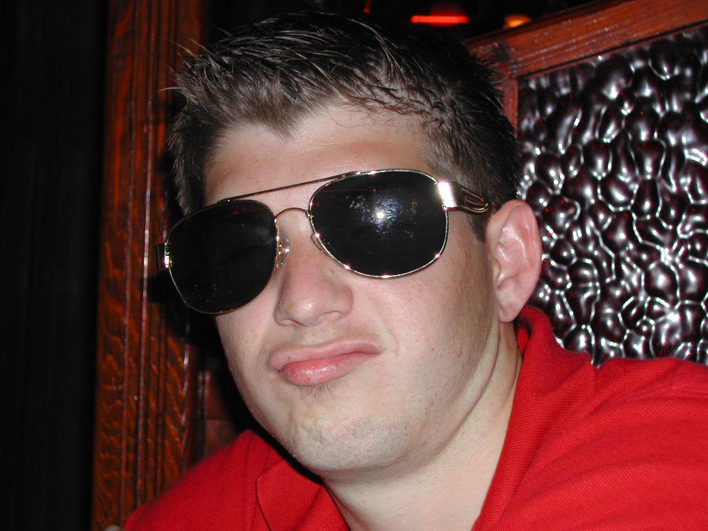A man in a red shirt and sunglasses makes a playful expression while sitting in a dimly lit setting.