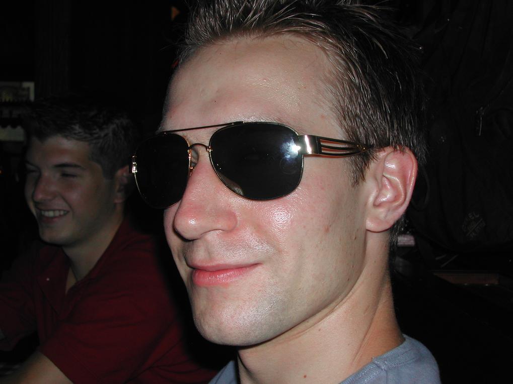 A man wearing dark sunglasses smiles slightly, with another person in the background also smiling.