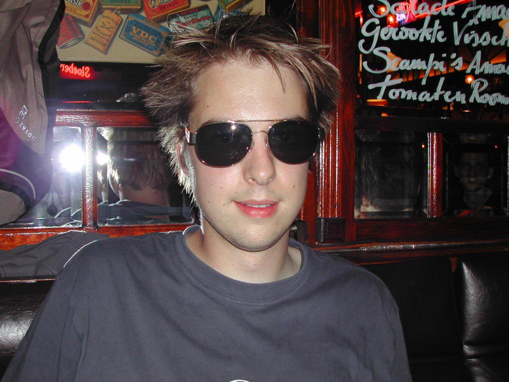 A man with spiky hair and sunglasses sits in a dimly lit bar with mirrors and handwritten menu text.