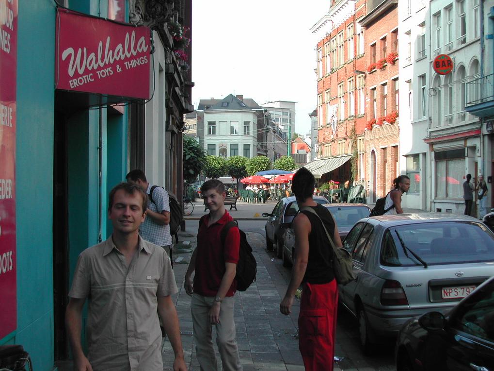 A group of young men stand on a city sidewalk near a shop with a red sign.