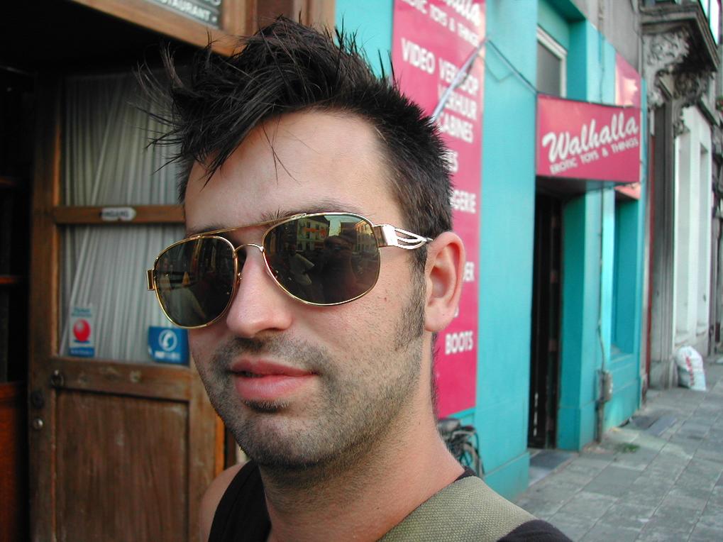 A man with spiky hair and sunglasses looks at the camera while standing on a city sidewalk.