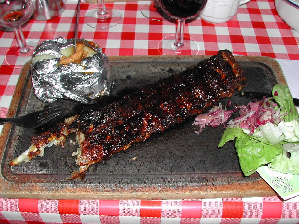 A plate with barbecued ribs, a baked potato with sauce, and a side salad on a checkered tablecloth.