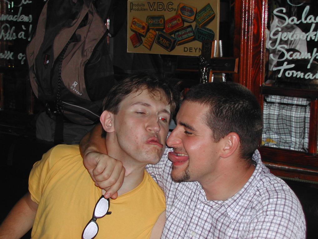 Two men sitting closely, one playfully sticking out his tongue while the other leans in with a relaxed expression.