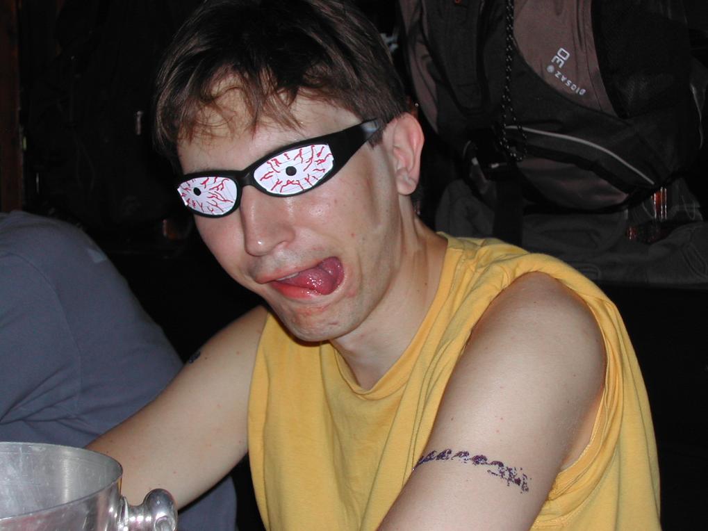 A man in a yellow shirt makes a playful face, wearing novelty glasses with cartoonish bloodshot eyes.