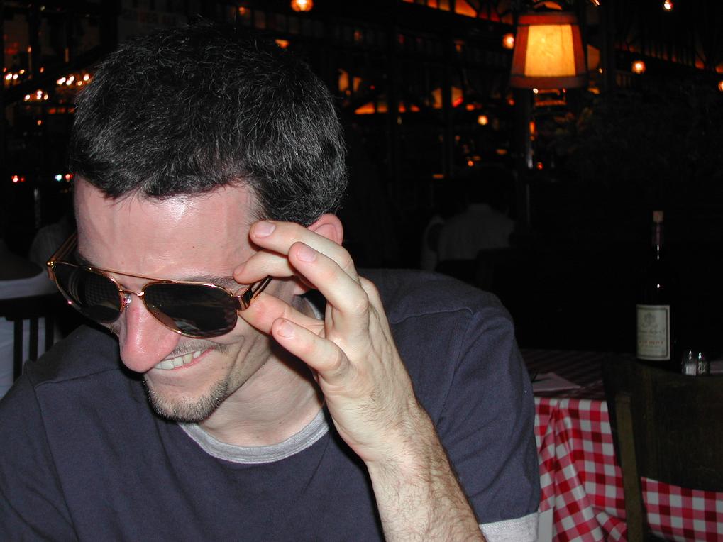 A man in a dark shirt laughs while adjusting his sunglasses at a restaurant with dim lighting.