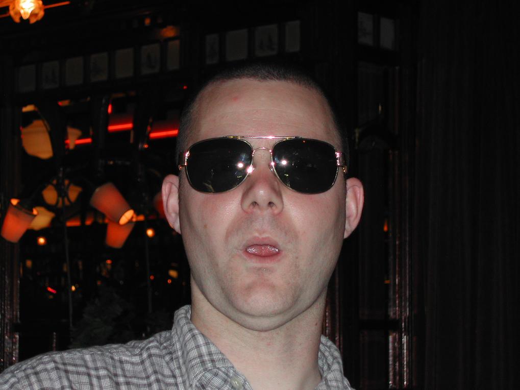 A man wearing sunglasses and a plaid shirt makes a playful expression with his mouth slightly open.
