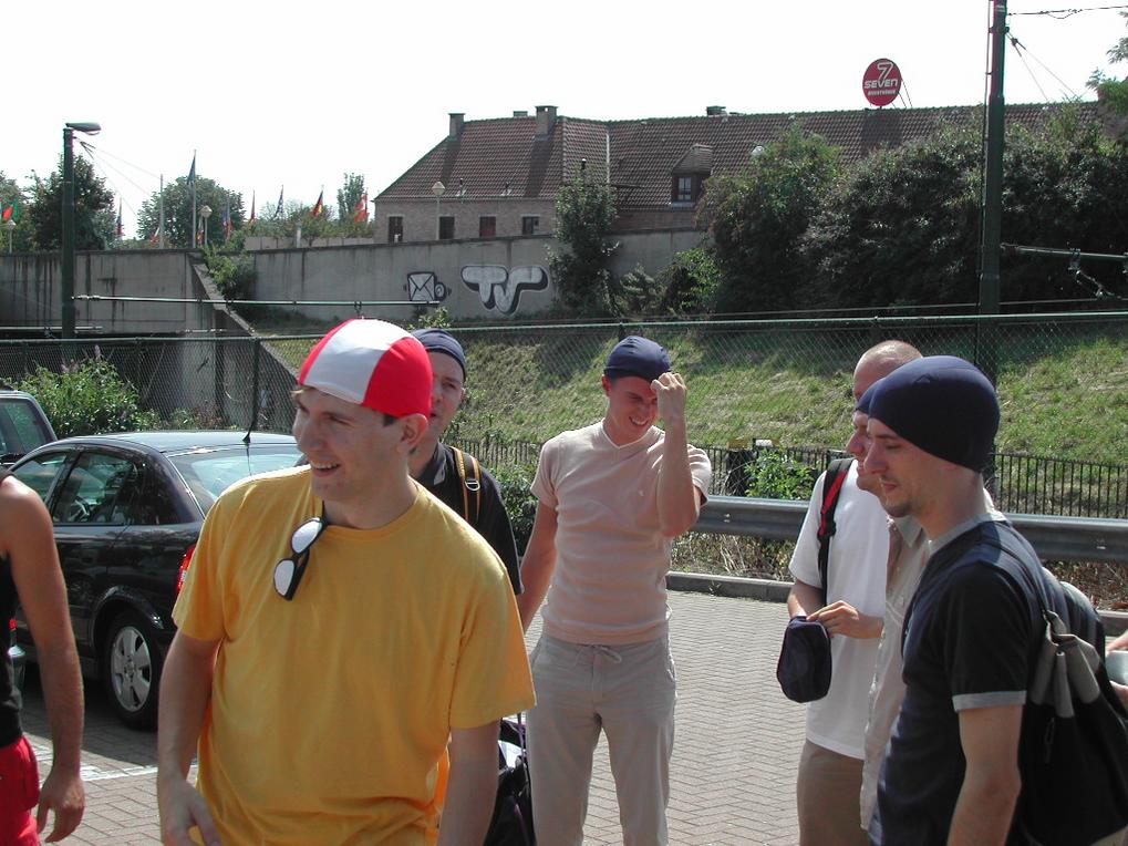 A group of men wearing casual clothes and hats stand together outside, smiling and chatting.
