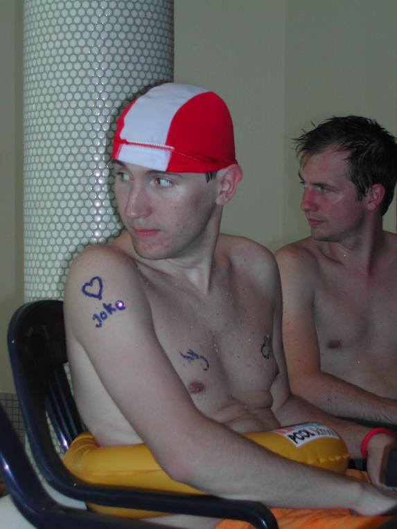 Two shirtless men sit near a pool, one wearing a red and white cap with writing on his arm.