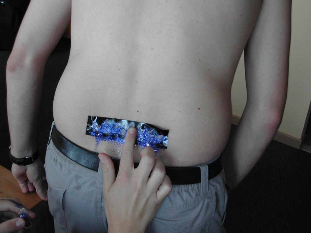 A person points at a small, glittery decoration taped to another person's lower back.