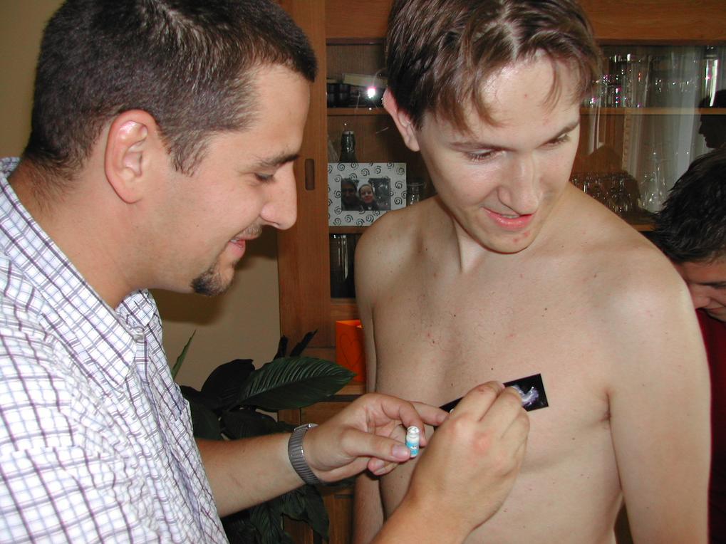 A man applies a temporary tattoo to a smiling, shirtless man's chest during a gathering.