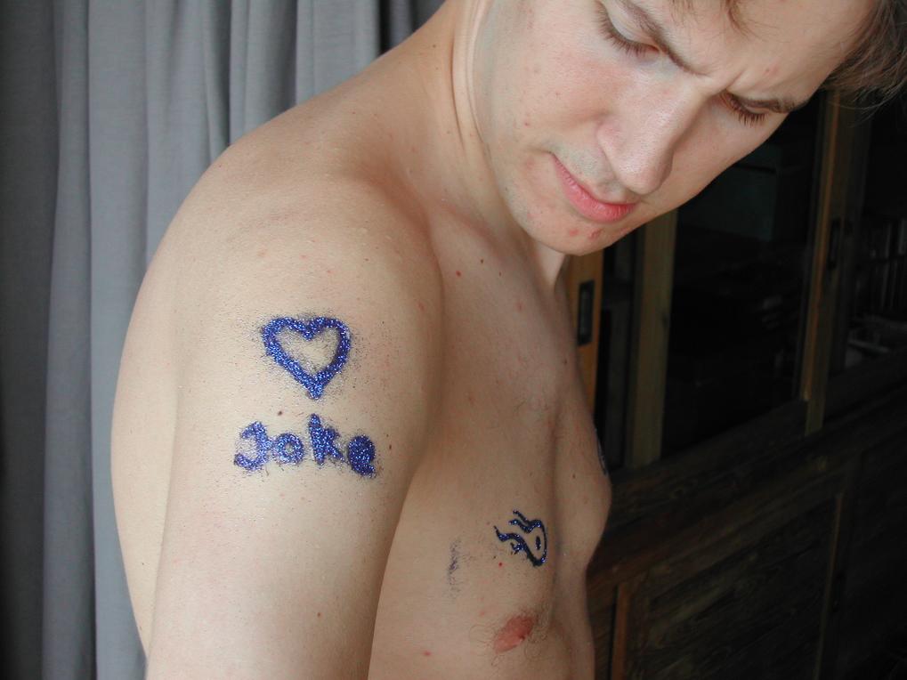 A shirtless man looks at his shoulder, which has a glittery temporary tattoo of a heart and the name "Joke.