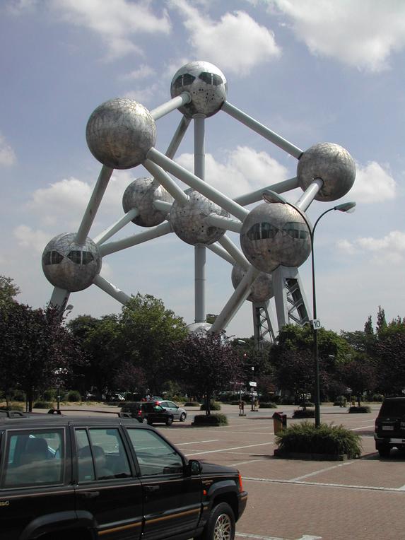 The Atomium, a large metallic structure with spherical nodes, stands in a public area with parked cars and trees.