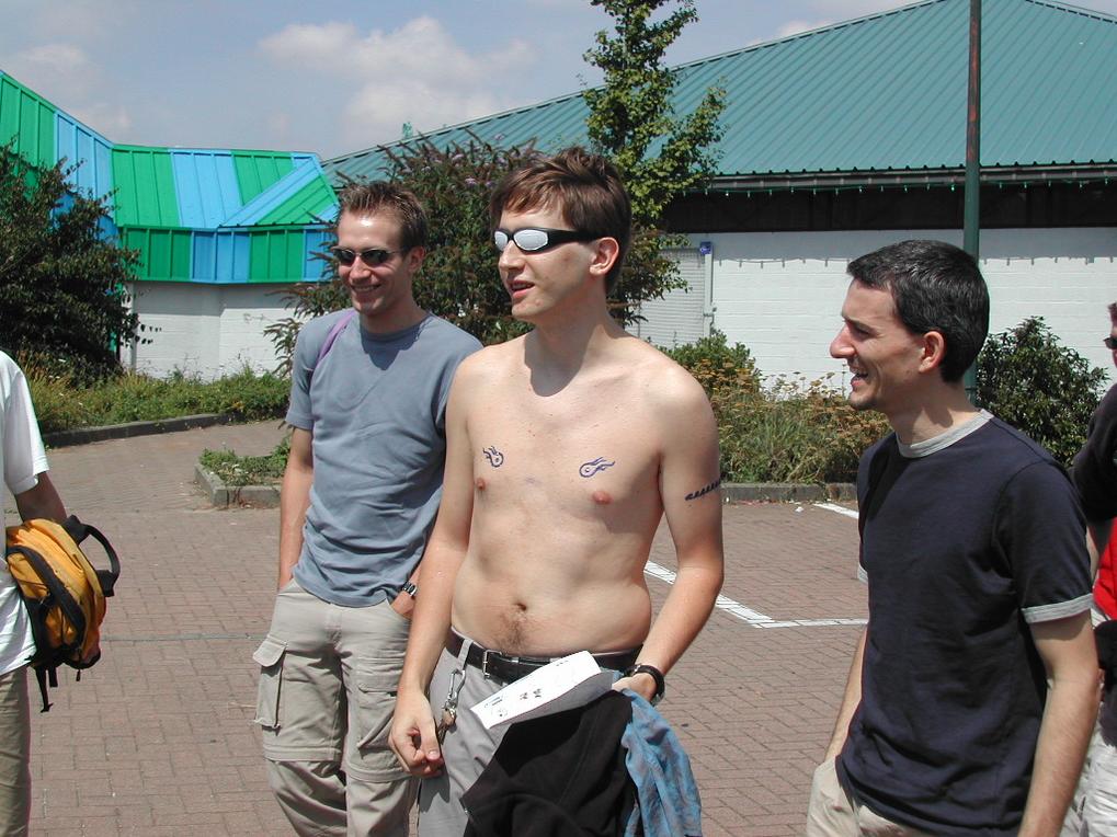 A shirtless man with sunglasses and temporary tattoos stands with friends, smiling and talking outdoors.