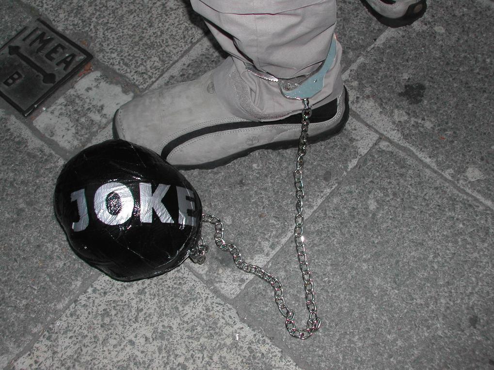 A person's ankle is shackled to a fake black ball labeled "JOKE" with a metal chain on the pavement.