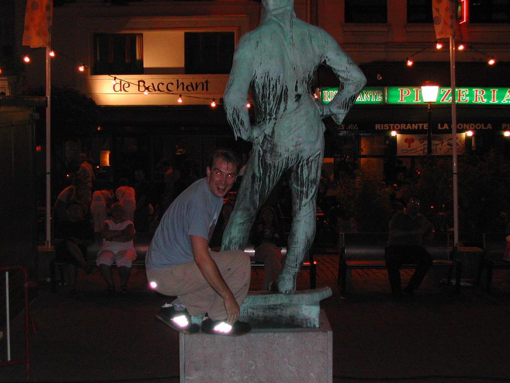 A man in a blue shirt crouches on a statue's pedestal, smiling at the camera during a night out.