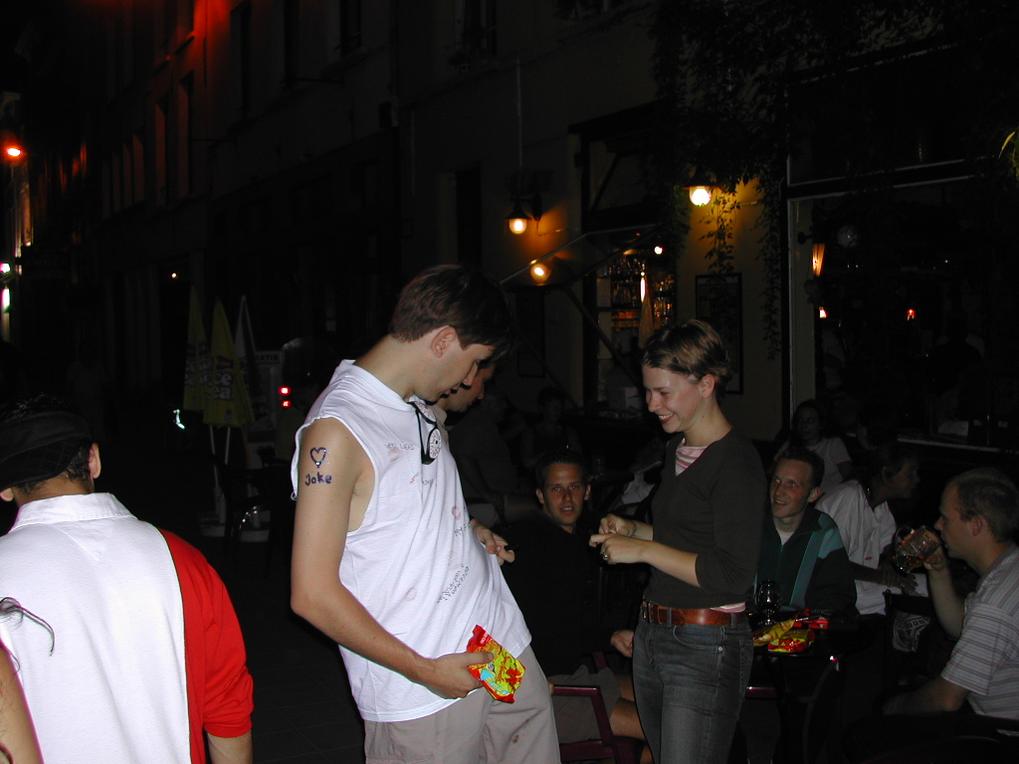 A man in a white shirt with writing on it interacts with a smiling woman at an outdoor bar.