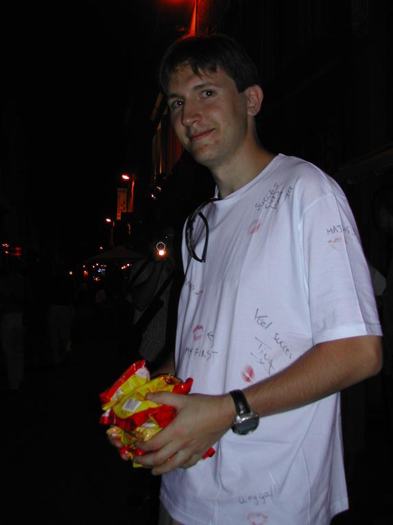 A man in a white shirt with writings and lipstick marks holds a bundle of snacks at night.