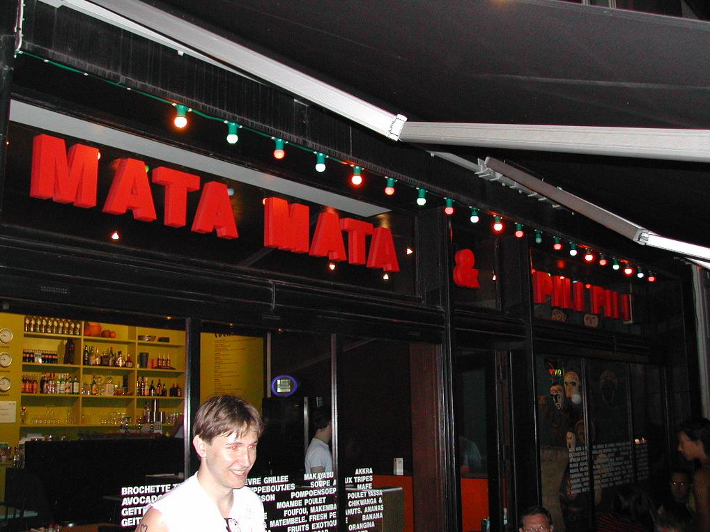 A man smiles outside a bar named "Mata Mata &amp; Pili Pili" during a nighttime pub crawl.