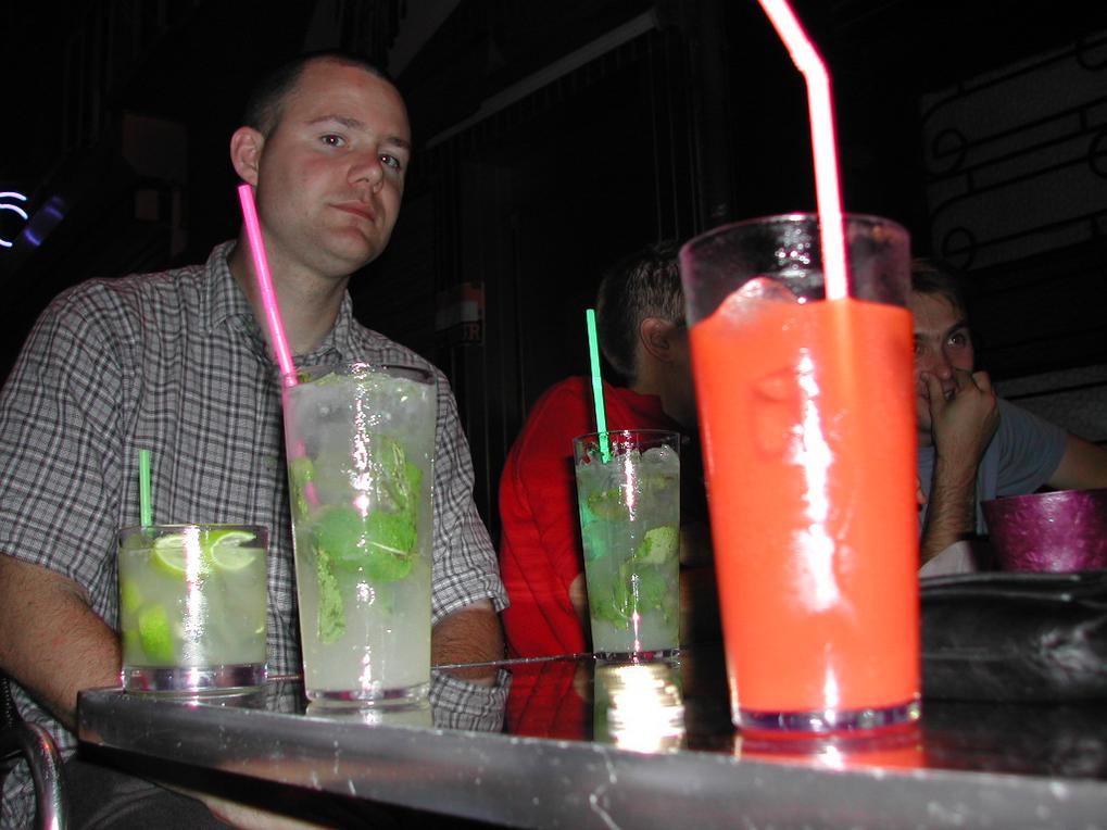 A man sits at a table with colorful cocktails, while others in the background engage in conversation.