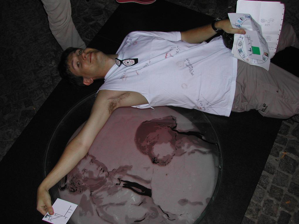 A man in a white shirt lies on the ground, holding a paper, with a glass artwork beneath him.
