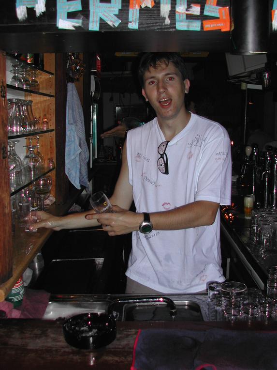 A man in a white shirt with signatures and lipstick marks stands behind a bar holding a glass.