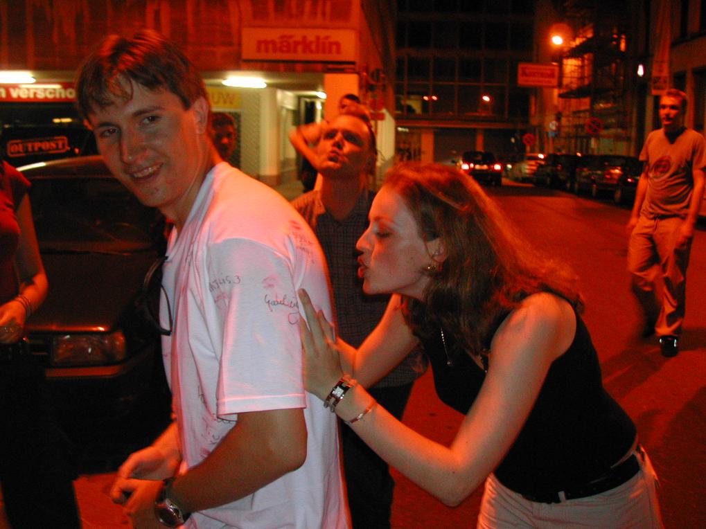A woman playfully kisses a man's shoulder as he smiles during a night out, wearing a signed white shirt.