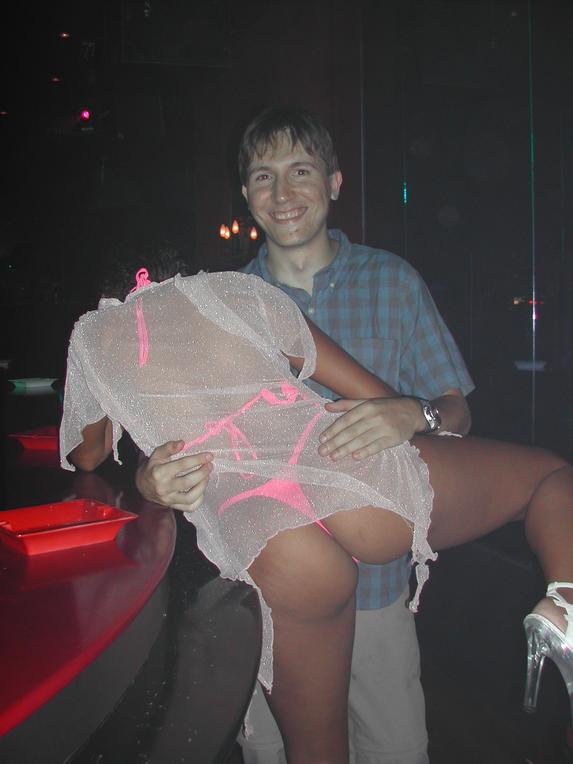 A man smiles at the camera while holding a woman dressed in sheer clothing and pink lingerie in a nightclub.