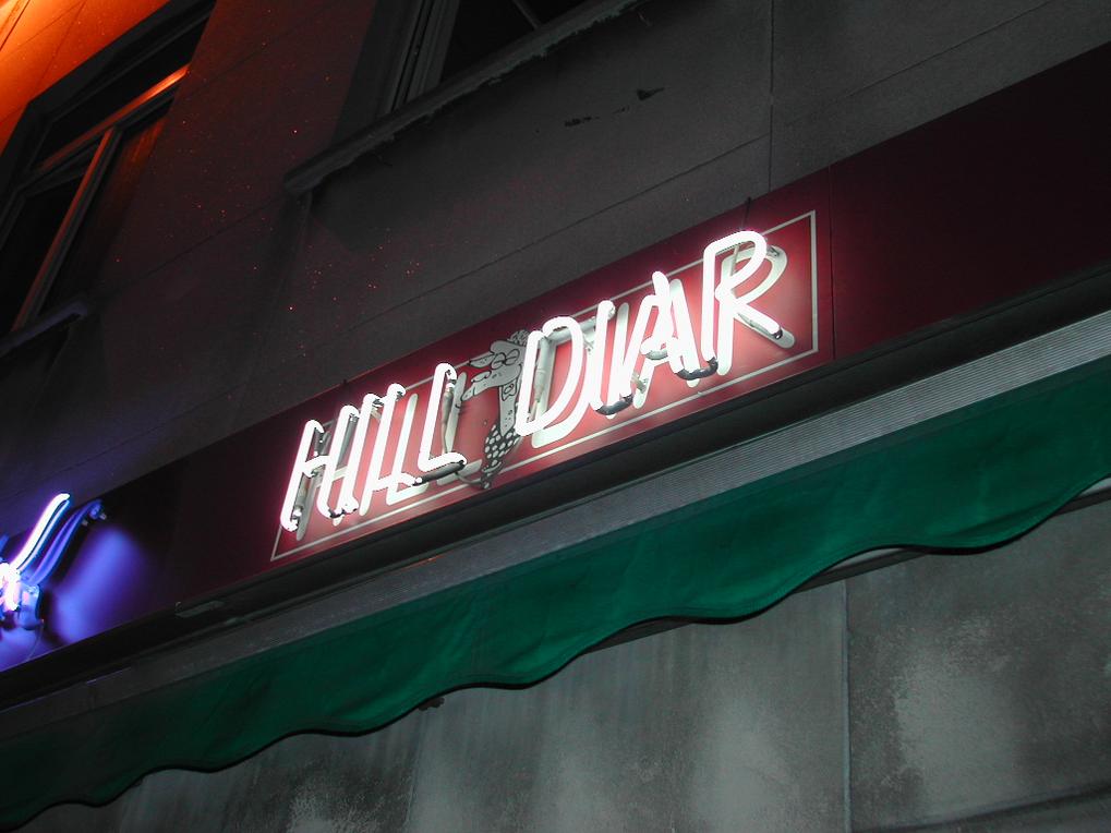 A neon sign reading "HILL DIAR" is mounted on a building, glowing at night above a green awning.