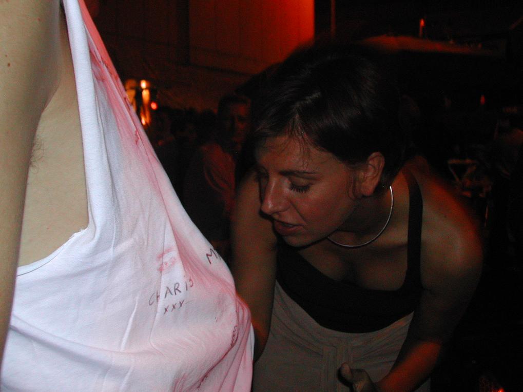 A woman leans in closely to examine or write on a man's white tank top covered in signatures and marks.