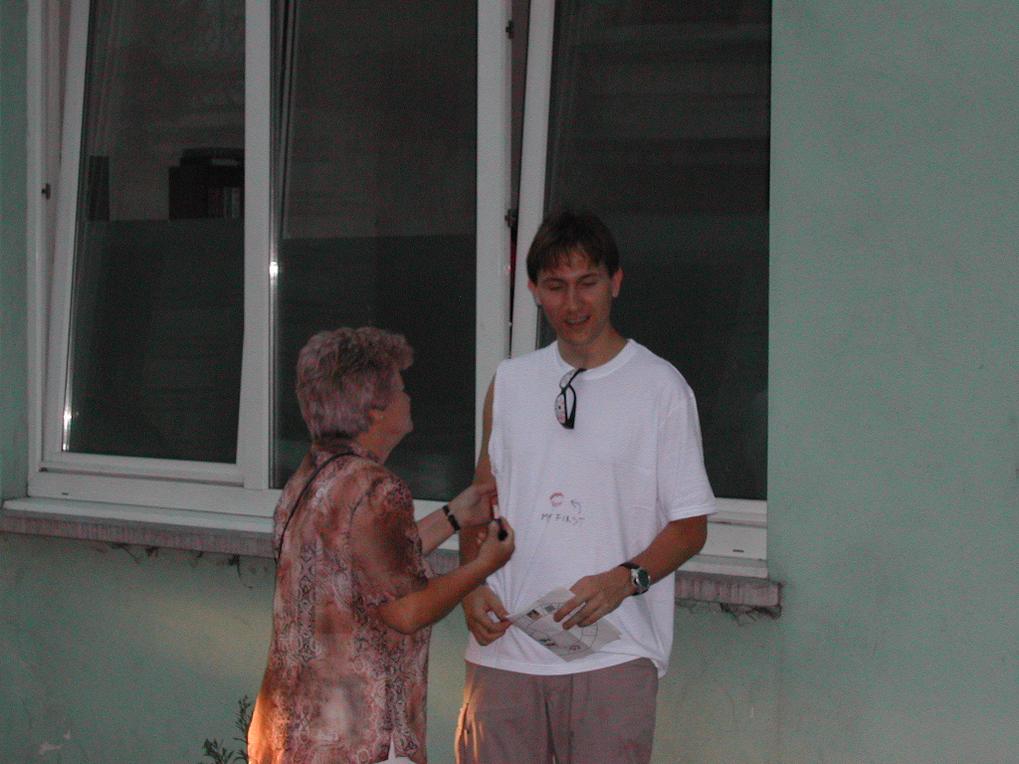 A woman and a man stand outside a building, talking while the man holds a piece of paper.
